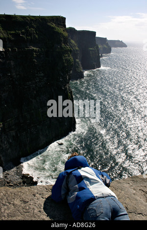 A young woman down on her stomach takes in the view of the Cliffs of Moher from the flat rock viewing place beyond the barrier Stock Photo