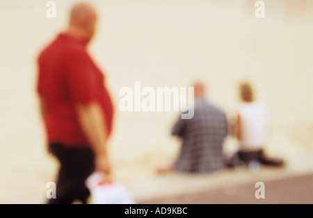 Defocussed man with large stomach in red shirt walking past and looking at a couple sitting on edge of seaside promenade Stock Photo