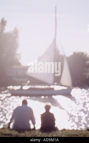 Impressionistic man and woman sitting on riverbank watching a yacht sailing past as they look into the sun Stock Photo
