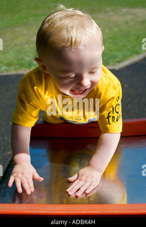 Vertical close up of a 16 month old baby boy looking at his reflection in a metallic slide, in the park on a beautiful sunny day Stock Photo