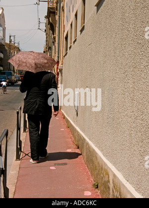 a lone male carrying umbrella as protection from sun walks away down  street Stock Photo