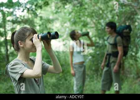 Three hikers standing in forest, young woman in foreground looking through binoculars Stock Photo