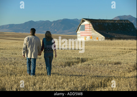 Couple walks through field of cut grass in Fall Colorado early morning horizontal toward old barn with american flag painted on Stock Photo