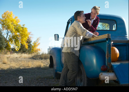 Middle aged female sitting in the bed of an old blue pickup truck feeding an apple to a man standing next to it Stock Photo