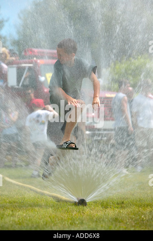 Boy leaps through fire hose spray at 4th of July celebration; firetruck and people in background Stock Photo