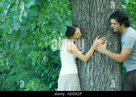 Man and woman leaning against either side of tree trunk, looking at each other, man holding woman's hand Stock Photo