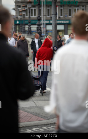 woman in red hoodie and blue trousers pulling a shopping wheeled trolley standing leaning against pedestrian crossing waiting on Stock Photo