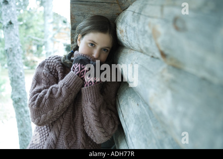 Teen girl leaning against wooden cabin, pulling turtle neck over mouth Stock Photo