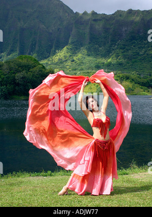 Belly dancer with red veil arms outstretched in tropical setting Stock Photo