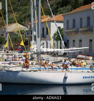 Row of moored yachts with people lounging onboard and trees and houses behind in Kioni Ithaca Island Greek Islands Greece Stock Photo
