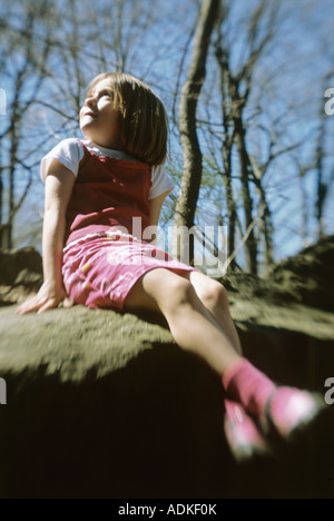 Little girl sitting on rock at park, looking upward Stock Photo