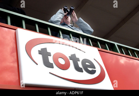 A racegoer studies form with binoculars over a Tote logo on a balcony at Lingfield Park racecourse,  Surrey. Stock Photo