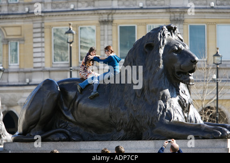 Tourists pose for photographs on lion statues in Trafalgar Square London UK Stock Photo