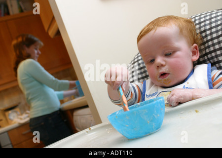A baby feeding himself in a high chair whilst his mother washes the dishes in the kitchen Stock Photo