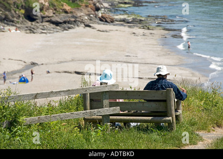 Pendower Beach at Gerrans Bay couple wearing sun hats relaxing on rustic bench overlooking quiet Cornish sandy beach near Veryan Cornwall England UK Stock Photo