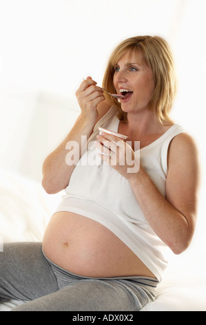 ATTRACTIVE HEALTHY YOUNG PREGNANT WOMAN SITTING ON BED AND EATING FRUIT YOGHURT Stock Photo