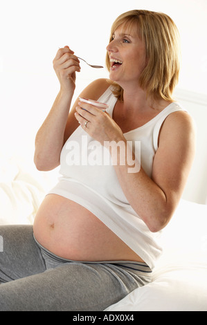 ATTRACTIVE HEALTHY YOUNG PREGNANT WOMAN SITTING ON BED AND EATING FRUIT YOGHURT Stock Photo