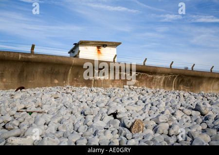 Concrete sea wall on the south Wales coast with pebbles piled up against it backed by a security fence old building and blue sky Stock Photo