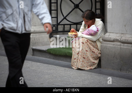 young gypsy beggar woman holding what looks like a baby wrapped in blanket eating chips in doorway as man walks past oconnell street dublin Stock Photo