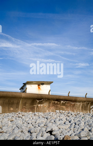 Concrete sea wall on the south Wales coast with pebbles piled up against it backed by a security fence old building and blue sky Stock Photo