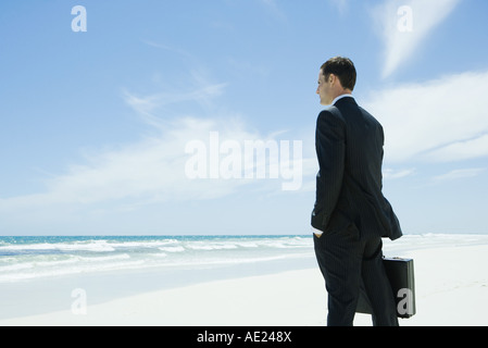 Businessman standing on beach, holding briefcase, facing ocean Stock Photo