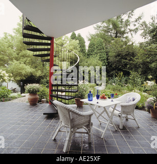 White wicker chairs and small table on slate paved patio below metal spiral staircase of modern house with lushly planted garden Stock Photo