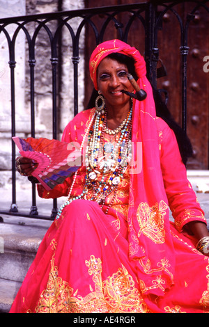 Cuban woman wearing flanboyant clothes in Old Havana She is wearing earings and many necklaces and is smoking a cigar on a pipe Stock Photo