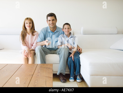 Father and two children sitting on sofa, pointing remote control Stock Photo
