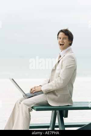 Businessman sitting on edge of picnic table at beach, using laptop Stock Photo