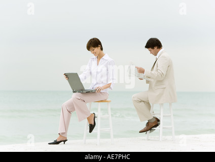 Businesspeople sitting on stools on beach, one using laptop, another reading newspaper Stock Photo