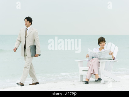 On beach, businessman carrying laptop, walking by woman reading newspaper in chair Stock Photo