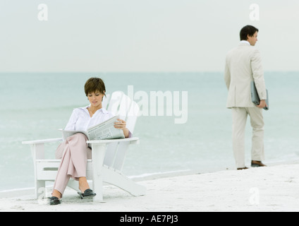 On beach, businessman carrying laptop, walking by woman reading newspaper in chair Stock Photo