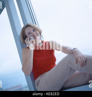 Businesswoman using mobile phone and sitting on windowsill Stock Photo
