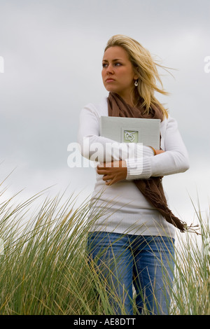 A beautiful blond haired blue eyed young woman stands amid long grass on a windswept headland clutching a book of memories Stock Photo