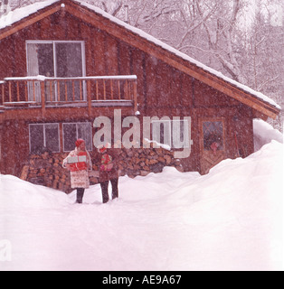 Christmas scene with couple leaving snow covered rustic mountain home carrying armloads of wrapped presents Stock Photo