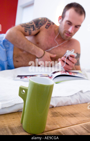 Tattooed man, barechested,  iPod player, lying on a bed, mug in foreground Stock Photo