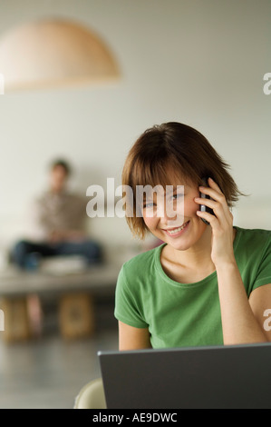 Smiling woman using laptop computer and mobile phone, man in the background Stock Photo