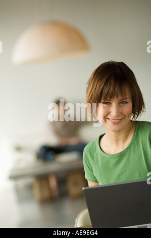 Smiling woman using laptop computer, man in the background Stock Photo