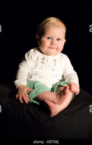 A full length portrait of a 6 month old baby girl sitting down with a black background. Stock Photo