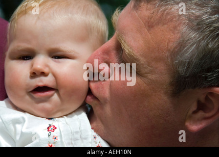 Horizontal close up portrait of six-month old baby girl pulling a disapproving funny face as her father kisses her on the cheek Stock Photo