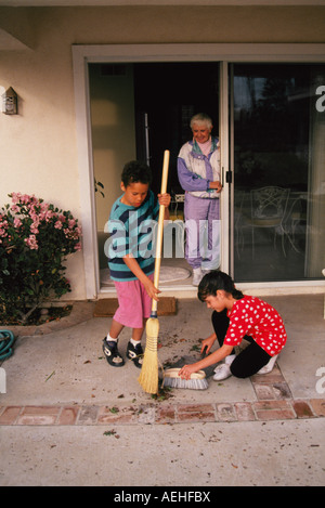 Diverse child helping another boy girl 6-8 year old older woman tidying sweeping sweeps sweep patio deck porch serving serve volunteer helps elderly Stock Photo