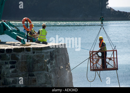 Mullion harbour wall entrance storm damage repairs by workmen using specialised drilling & access cage suspended over sea Mullion Cornwall England UK Stock Photo