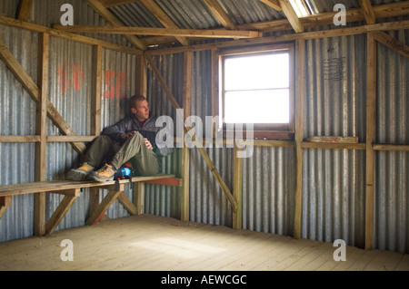 male hiker sits on bench waiting out summer snow storm in mountain shelter, Nelson Lakes national park, New Zealand Stock Photo