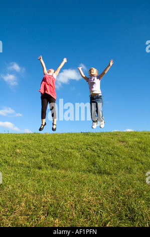 Two Girls Jumping On Top Of A Hill Against Blue Sky. Stock Photo