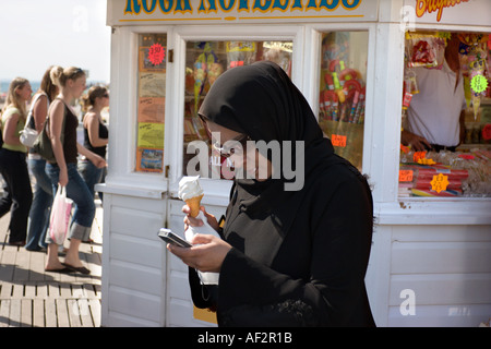 A Muslim woman eats ice cream and checks her mobile phone on the pier at Brighton Sussex England Stock Photo