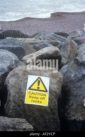 Sea defence rocks with pink shingle beach and grey sea beyond with sign fixed to rock stating Caution Keep off rocks Stock Photo