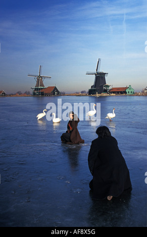 Netherlands Zaanse Schans Japanese tourists and swans on frozen river with windmills in background Stock Photo