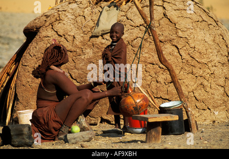 Himba woman and child making cream in calabash outside their dome-shaped mud hut Kunene River, Kaokoveld; Namibia Stock Photo