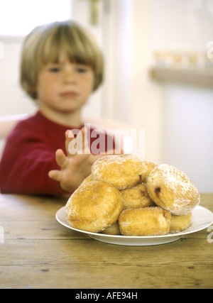 Young boy sitting down at a kitchen table reaching out for a doughnut Stock Photo