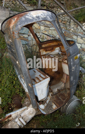 hulk of a rusted out abandoned three wheeled truck sitting in weeds in front of a stone wall on a beer keg Stock Photo